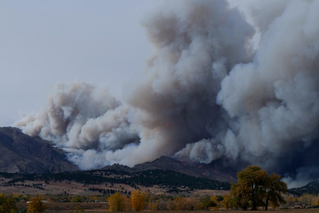 Aerial view of a wildfire burning near a highway, with smoke rising over prairie landscape and vehicles evacuating west.