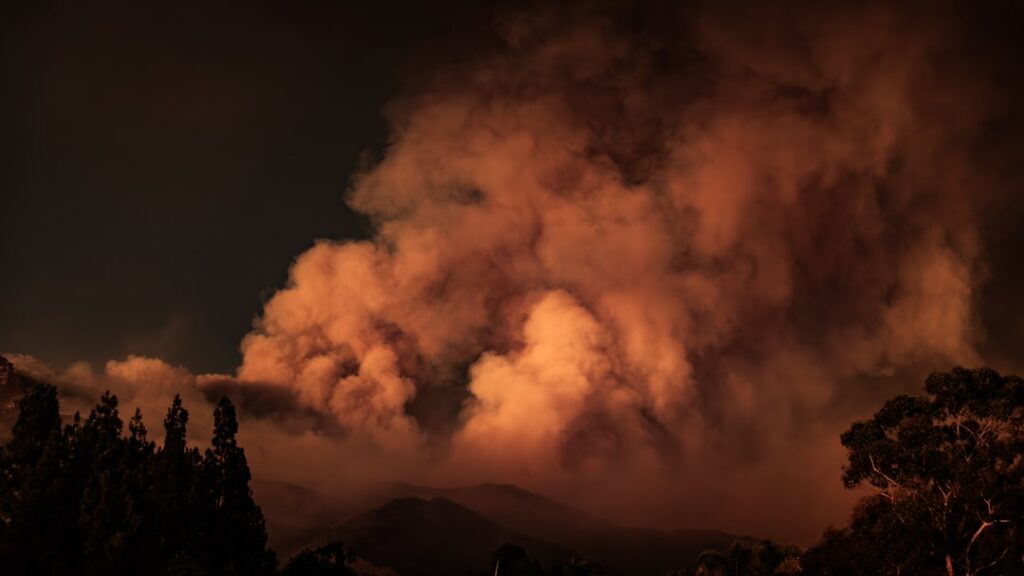 Orange flames race up dry hills toward Castaic Lake as smoke towers over fleeing traffic and emergency vehicles.