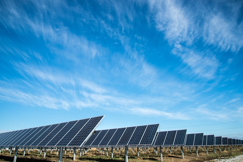 Rows of solar panels stretch across a plain as wind turbines spin on distant hills under a bright sky.