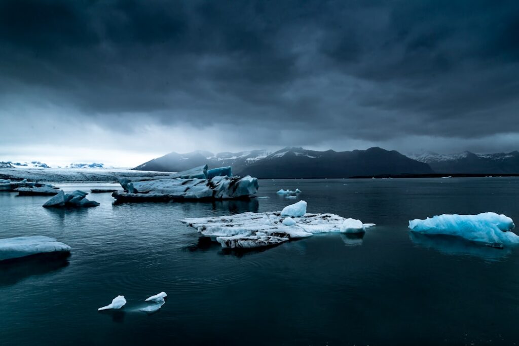 A massive Antarctic ice sheet stretches across the horizon under a pale blue sky, with penguins gathered on a rocky outcrop in the foreground.
