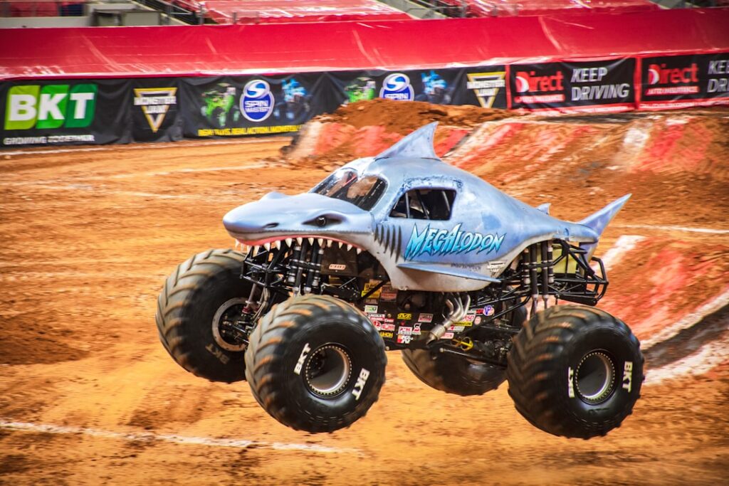 A large monster truck with oversized tires sits on a dirt track, surrounded by crowd barriers and spectators.