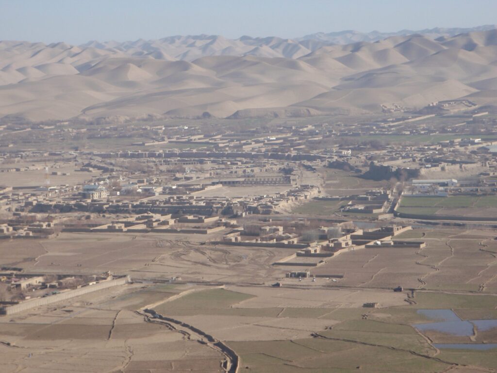 An overturned passenger bus lies on a rural road in Badghis Province, Afghanistan, with emergency responders nearby.