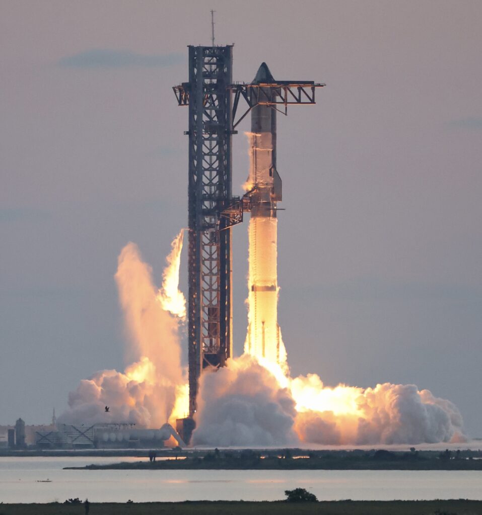 SpaceX Starship rocket lifts off from launch pad with exhaust plumes and bright flames against a clear sky.